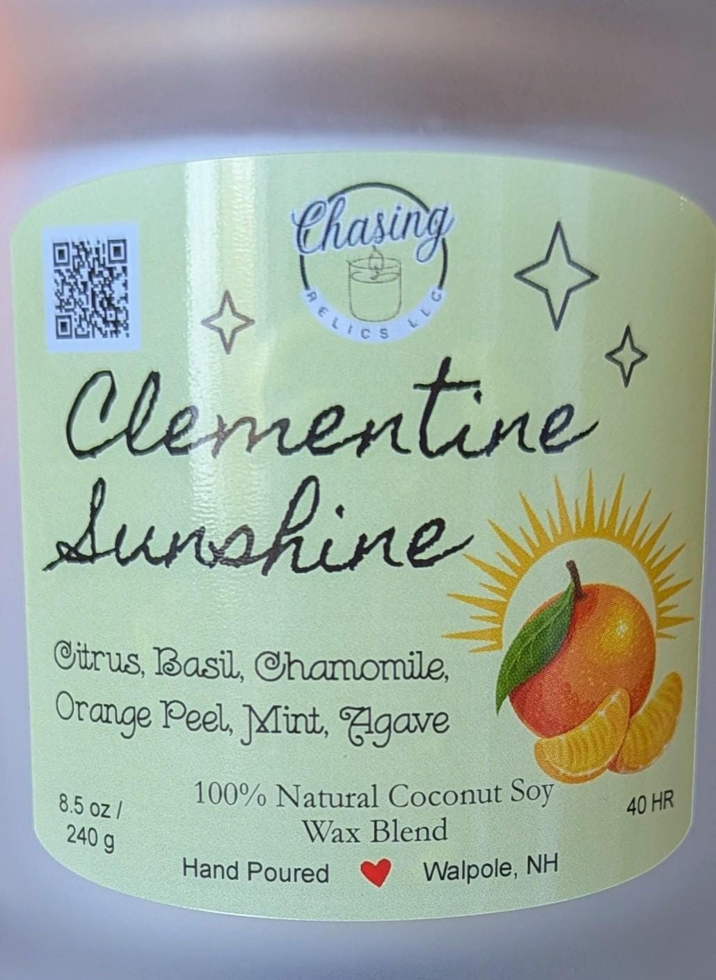A white candle jar with yellow label. The label has a QR code, the words 'Chasing Relics LLC', 'Clementine Sunshine', 'Citrus, Basil, Chamomile, Orange Peel, Mint, Agave', '100% Natural Coconut Soy Wax Blend', '8.5 oz/ 240 g', 'Hand Poured', and 'Walpole, NH'. The label also features a red heart and a picture of a clementine and a sun. The candle is 40 hours.