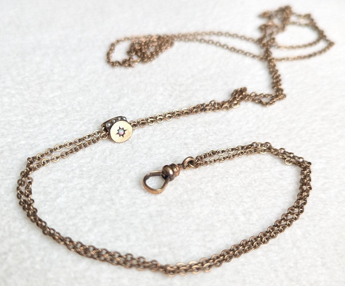 May include: A vintage gold-toned necklace with a delicate chain and a circular pendant. The pendant features a small, light-colored stone at its center, surrounded by tiny embellishments. The necklace has a clasp at the end and is displayed on a white surface.