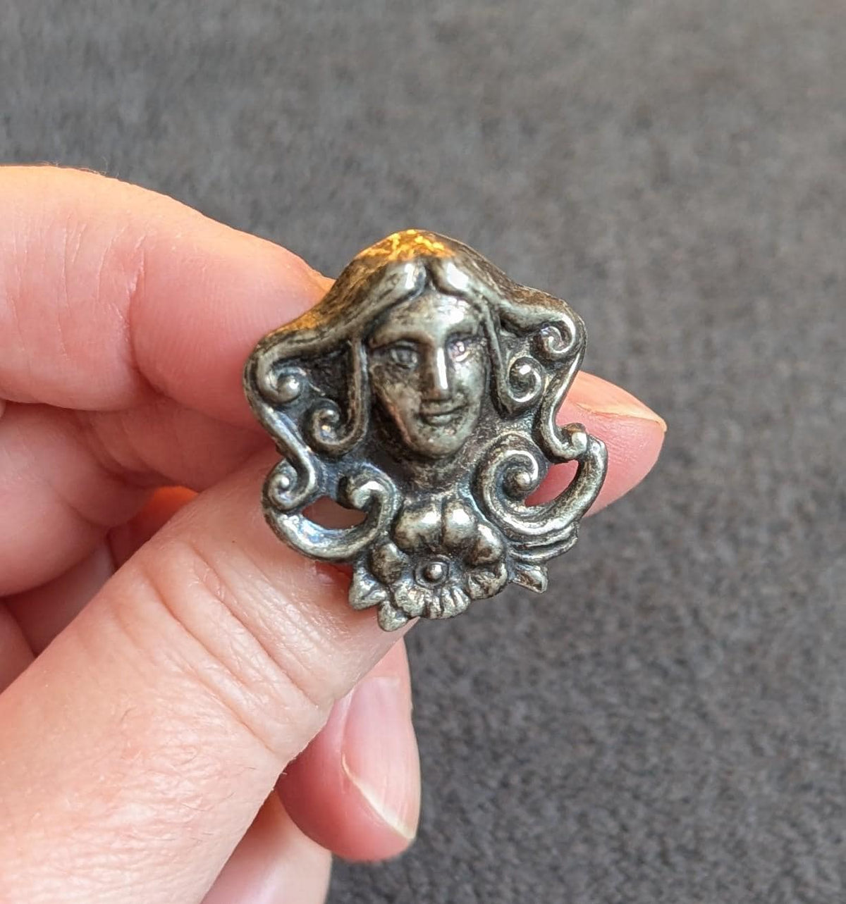 May include: A silver-toned, antique-style button or decorative element featuring a detailed face with flowing hair and floral accents. The design has an aged appearance, with intricate details and a vintage aesthetic.