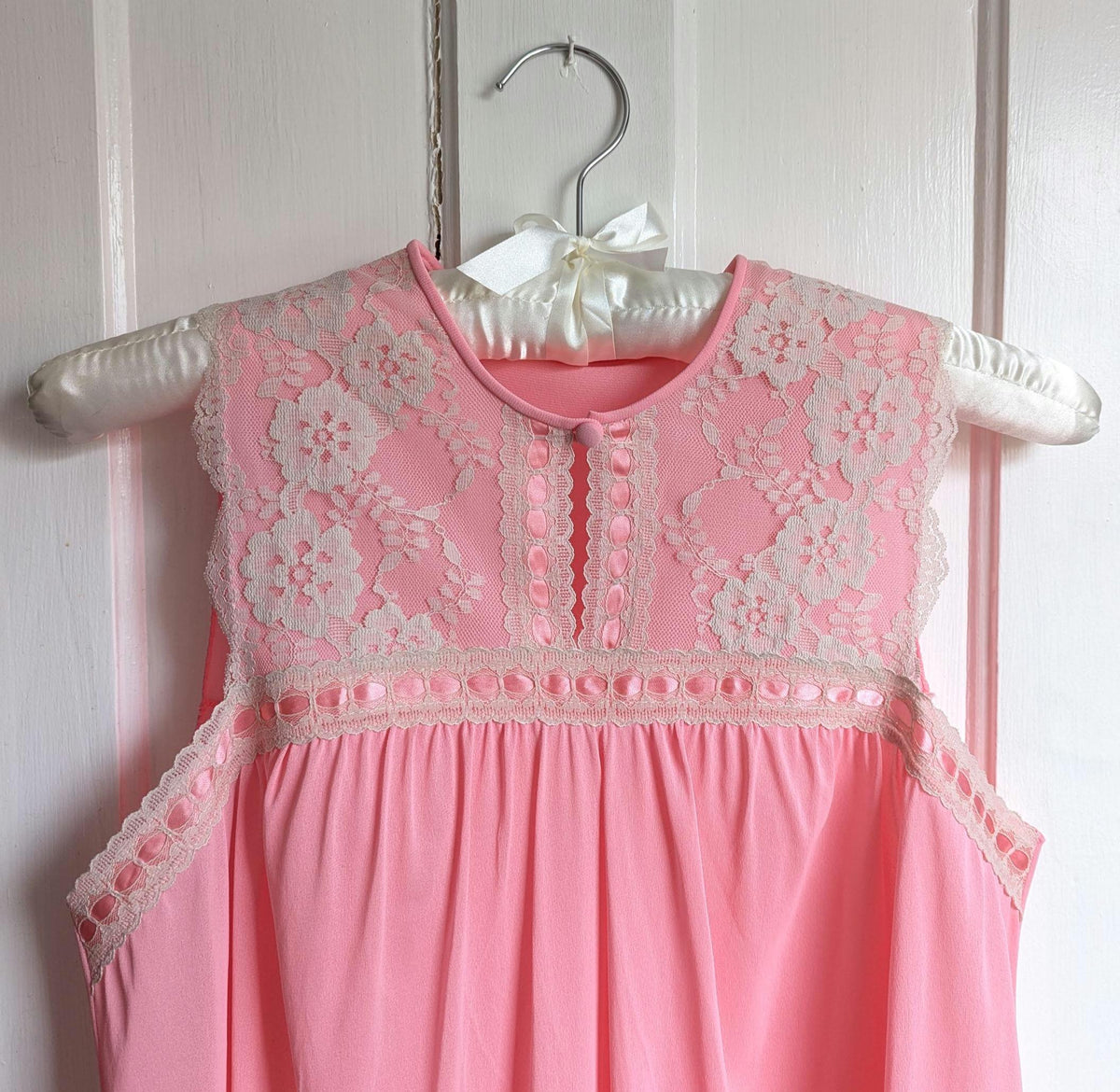 May include: A vintage pink nightgown with a floral lace yoke and trim. The nightgown is sleeveless and hangs on a satin hanger with a bow. The lace detailing is cream-colored, and the fabric has a soft, flowing appearance.