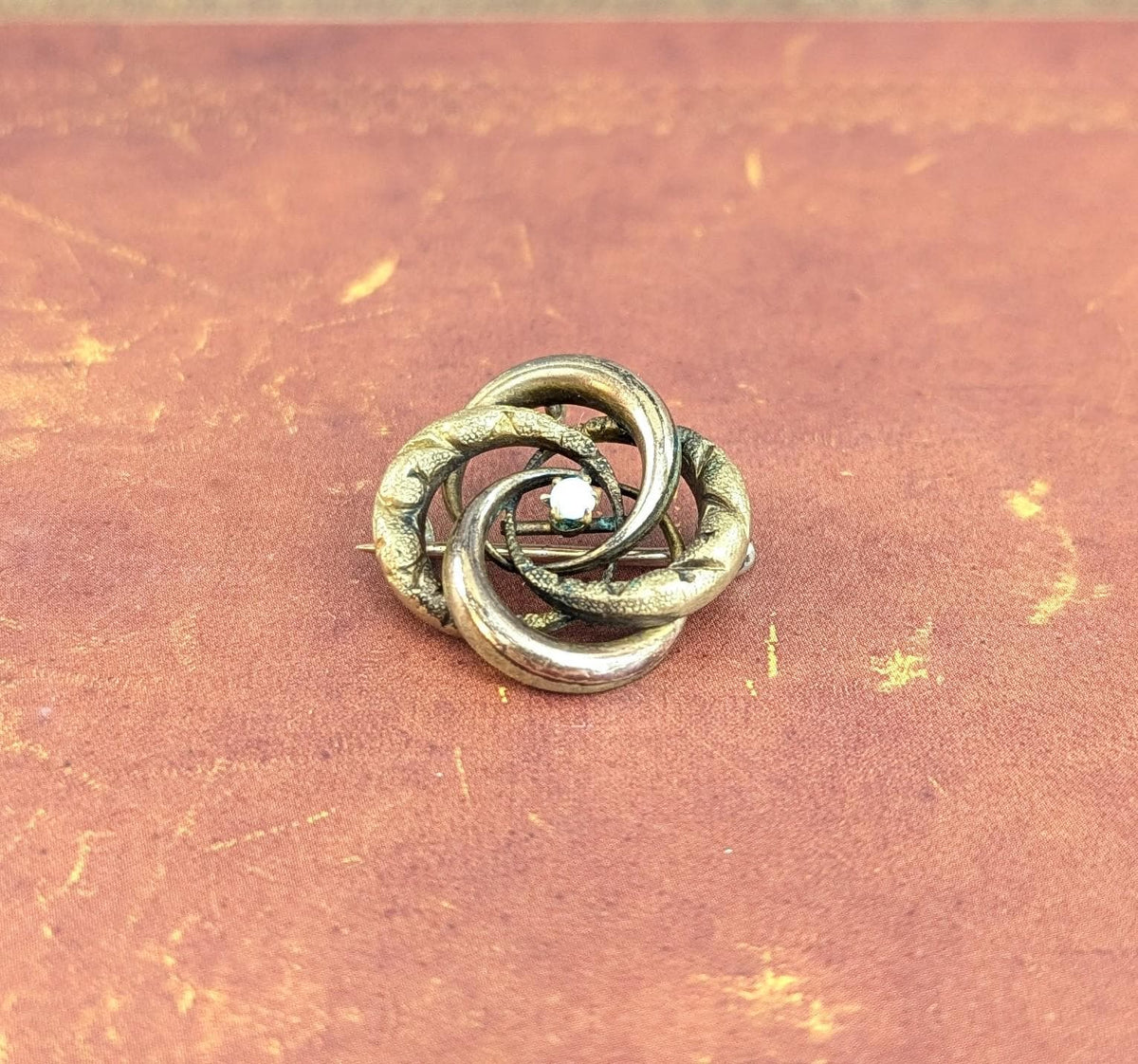 May include: A vintage silver-toned brooch with an intertwined knot design. The brooch features three interlocking loops with textured detailing and a small, central, clear gemstone. The brooch is set against a reddish-brown background.